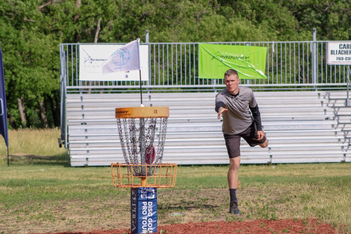 A disc golfer following through on a putt toward a chain basket on a grass course