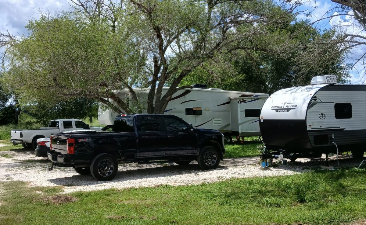 A pickup and travel trailer parked side by side on a wide gravel RV site under mature trees at The Texas Rose RV Park in Beeville, TX