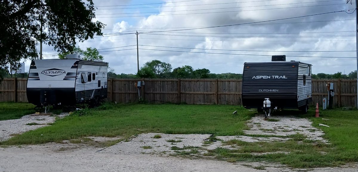 Two travel trailers on gravel pads with electric hookup boxes along the fence line at The Texas Rose RV Park in Beeville, TX